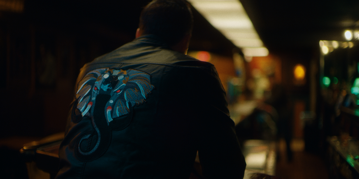 Moody cinematic narrative scene featuring a man seated at a bar wearing an embroidered jacket, filmed by Hiatt Films