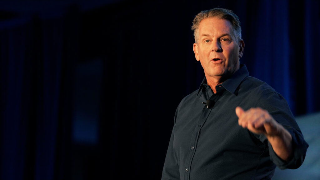 Male speaker presenting on stage with microphone and dark background